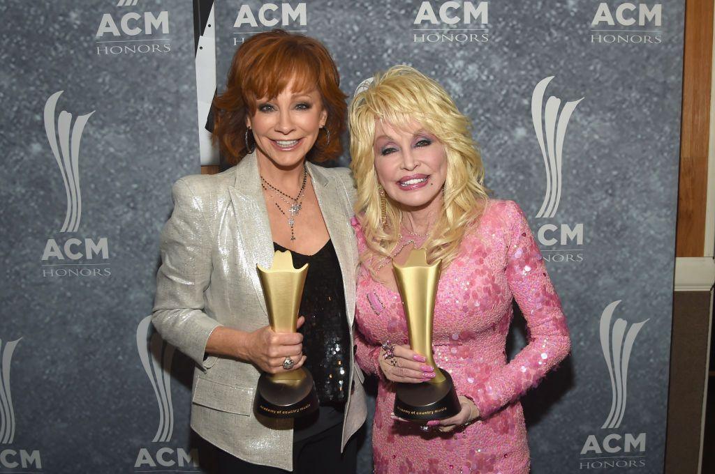 Reba and Dolly are pictures holding up two golden awards on a red carpet.