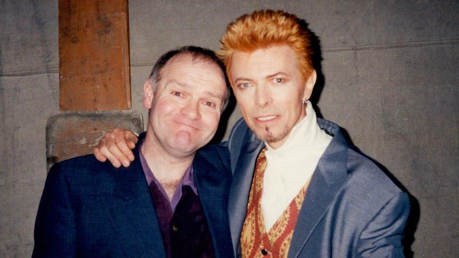 Two men are looking into a camera and smiling for a head and shoulders shot. The man on the left has short brown hair and is wearing a blue blazer and purple shirt unbuttoned a bit. On the right is singer David Bowie, who has orange hair spiked up, short stubble on hic chin, is wearing a blue jacket and patterned red and yellow waistcoat with a white top under it. He has his arm around the man next to him.