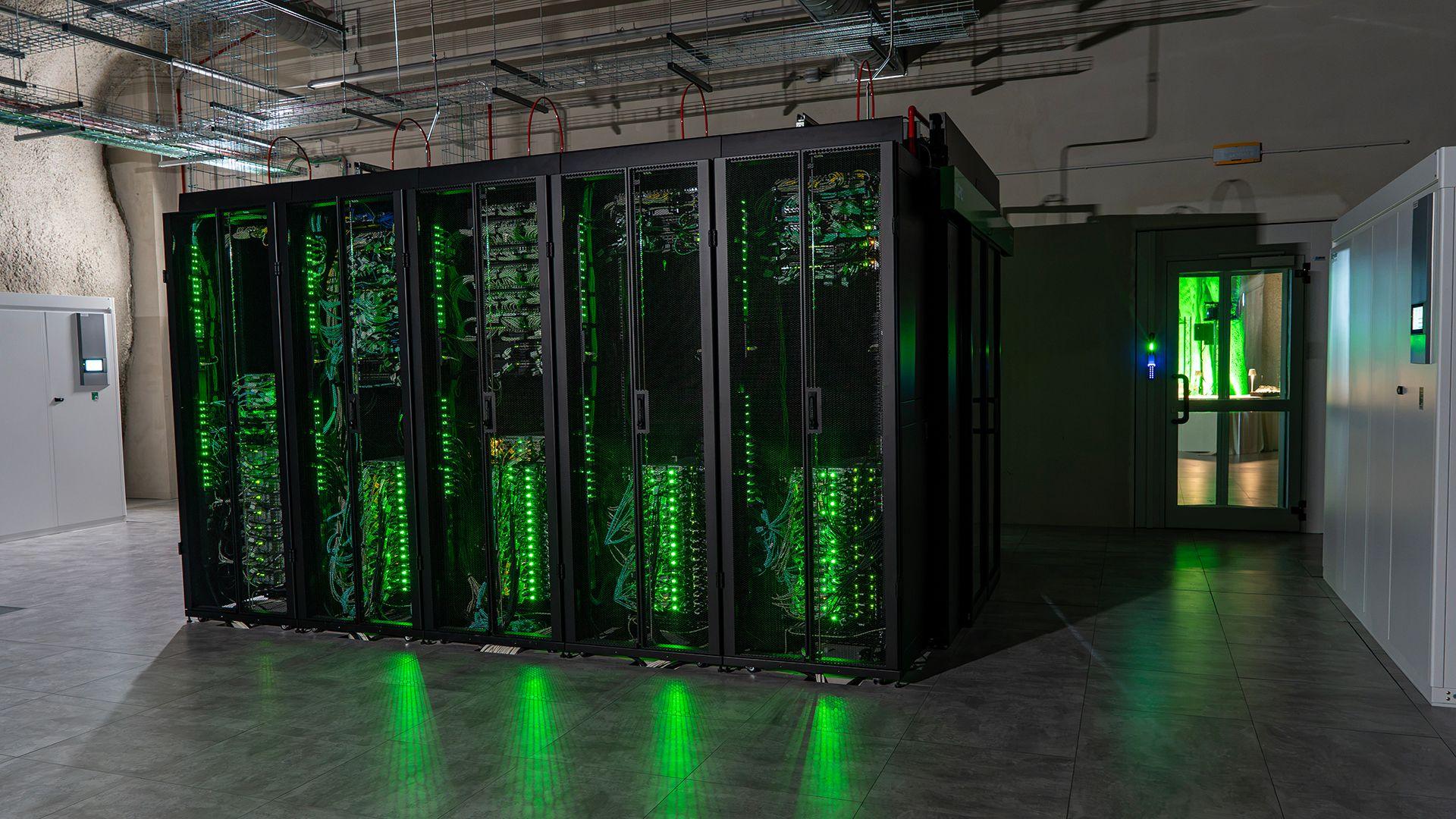 Green lights shine from a row of data servers in cabinets, reflecting on the high-gloss floor of the underground cavern. Metal trunking is attached to the ceiling above the cabinets, while to the right a door leads into a lighter room.