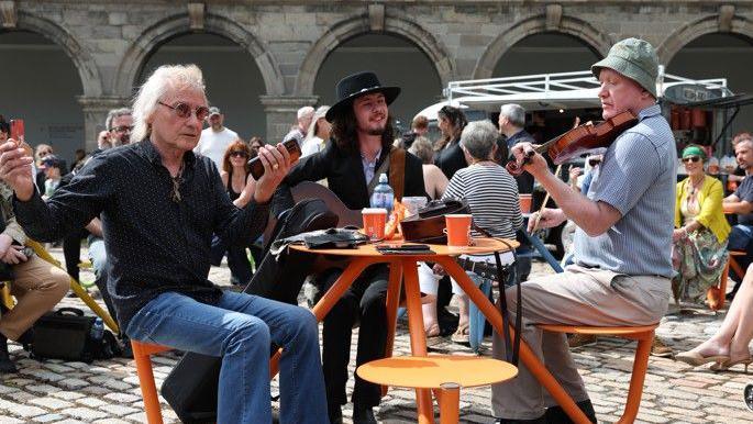 Three men sit at a table in the foreground of the picture. One man, to the right of the frame is playing a violin. He is wearing a light blue shirt, cream trousers and a green hat. He is facing towards the table. In teh middle of the table is a man with long dark hair and a short beard. He is wearing a black jacket and the strap of the guitar, he is playing, is visible. A third man faces towards the left hand side of the table. He is wearing a dark shirt and blue jeans. He has curly blonde or grey hair and sun glasses. He is holding a brown case in his hand. Other people are sitting in the background, listening to the music.