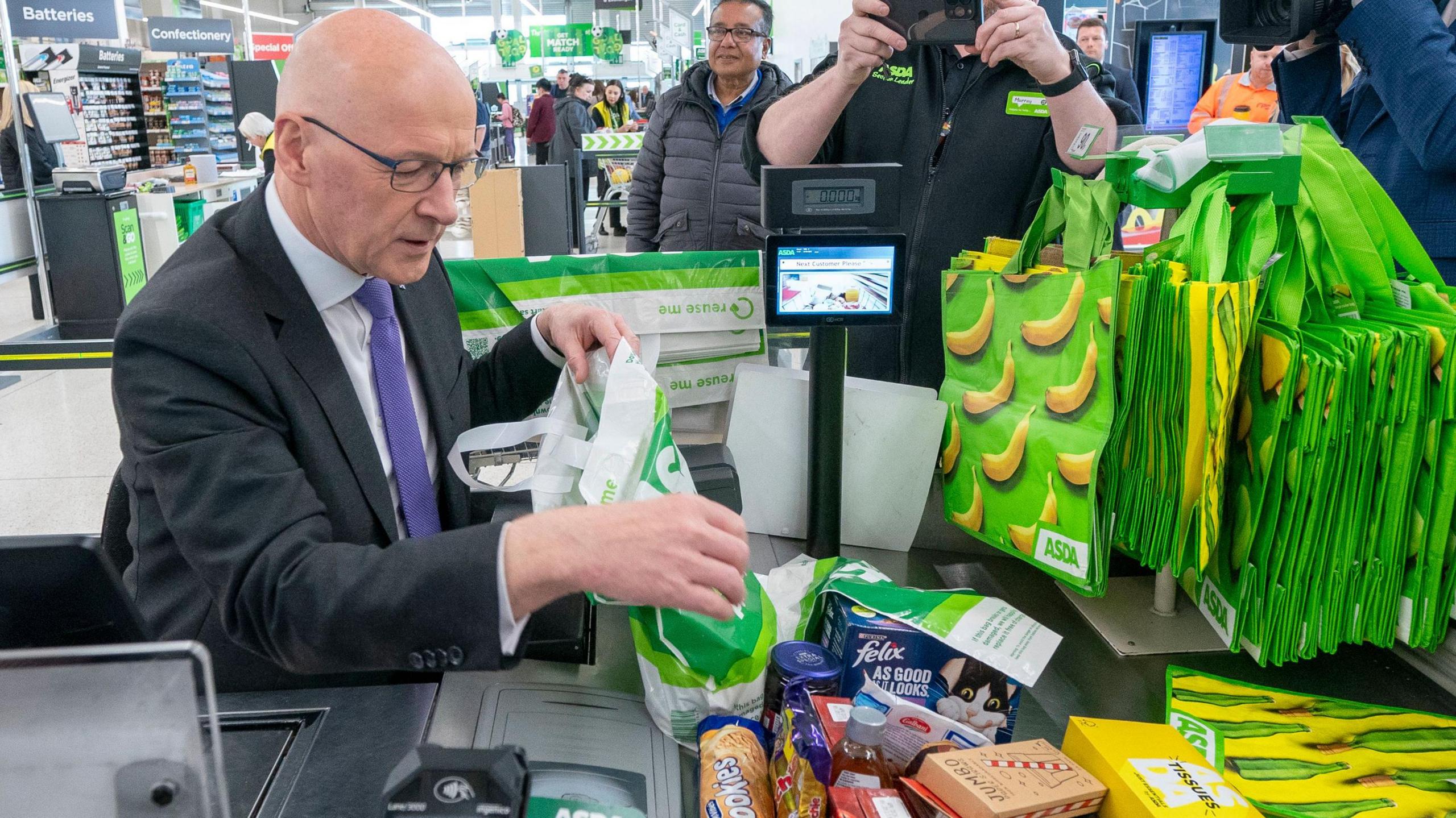 John Swinney is sitting at an Asda checkout where he is wearing a dark suit, white shirt and purple tie. He is bagging a pile of shopping as photographers and camera people capture the image.