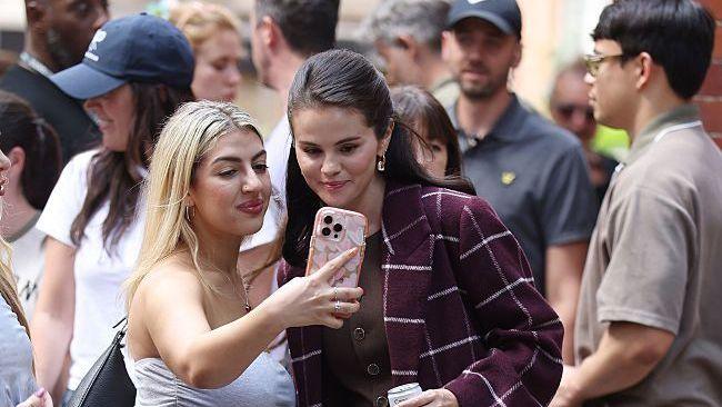 Selena Gomez in a red checked coat next to a fan who is taking a selfie of them both together.