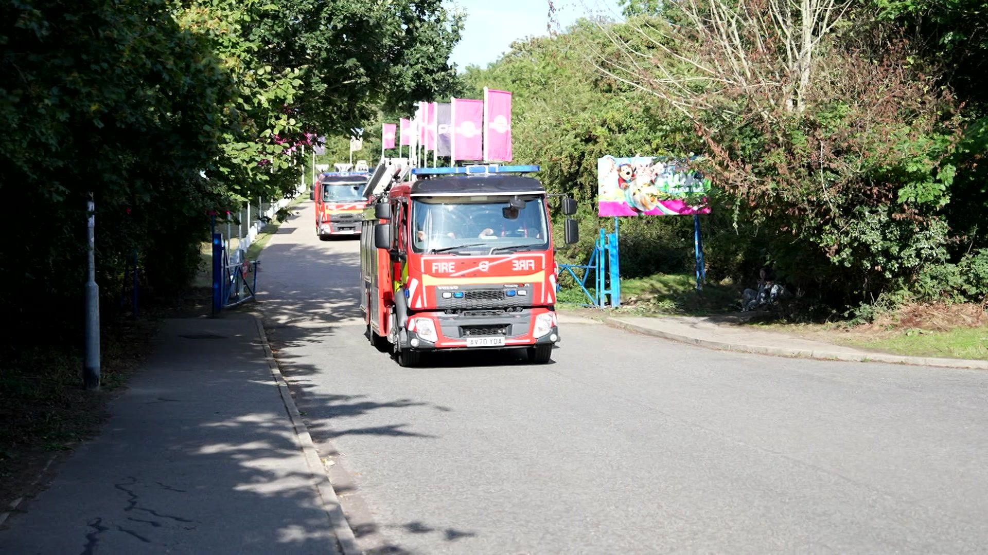 A red fire engine drives on a road, towards the camera, with another fire engine behind it. A sign, with animated images of animals printed on it, is fixed to blue posts on the right-hand side. There are green trees lining the road. It is sunny.