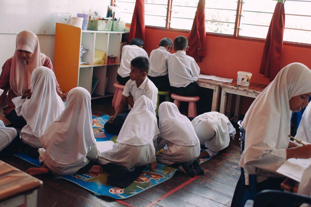 A classroom in Penang, 2017, with Rohingya children in white school uniforms working on lessons. Some sit together on a colourful mat writing and reading, while others sit on stools at a table beneath large windows with red curtains. Shelves with books and supplies line one side of the room, and a child in the foreground writes in a notebook.