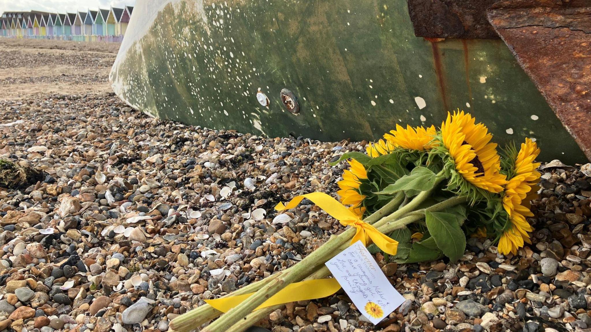 A bouquet of yellow sunflowers with a handwritten note attached laid on the pebbly beach. A row of colourful beach huts are pictured in the background.