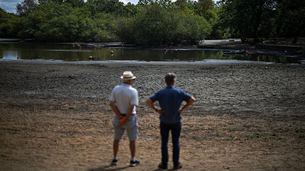 Two men standing on the dry bed of a reservoir looking at the little water remaining