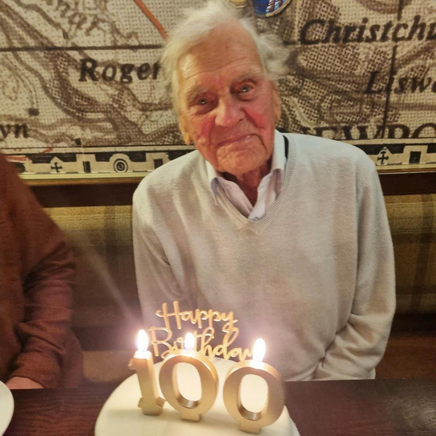 A man smiling at the camera. There is a cake in front of him with candles that say Happy Birthday 100.