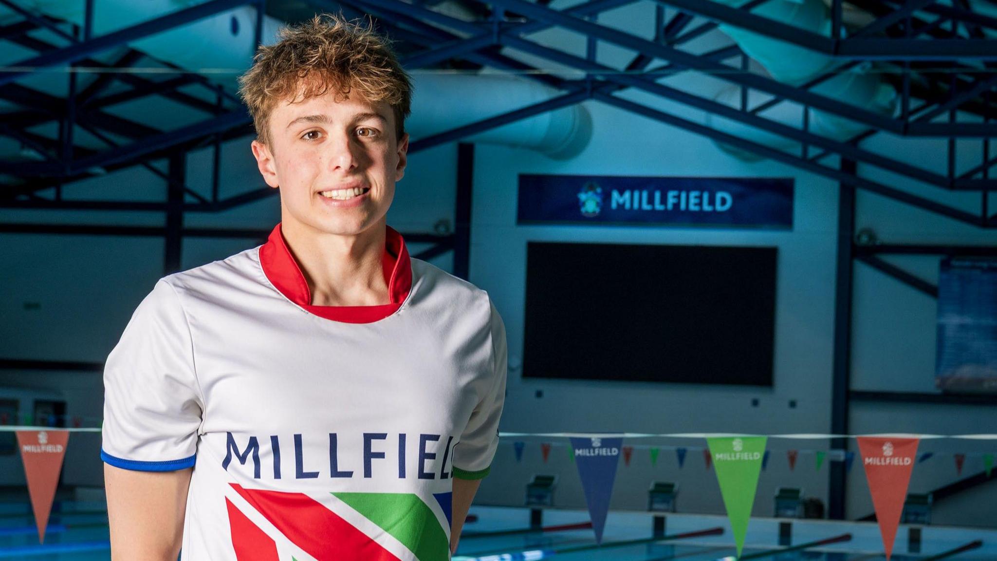 Filip Nowacki standing beside an indoor swimming pool, wearing a white Millfield sports shirt with a colourful flag design. Swimming lanes and Millfield banners are visible in the background.