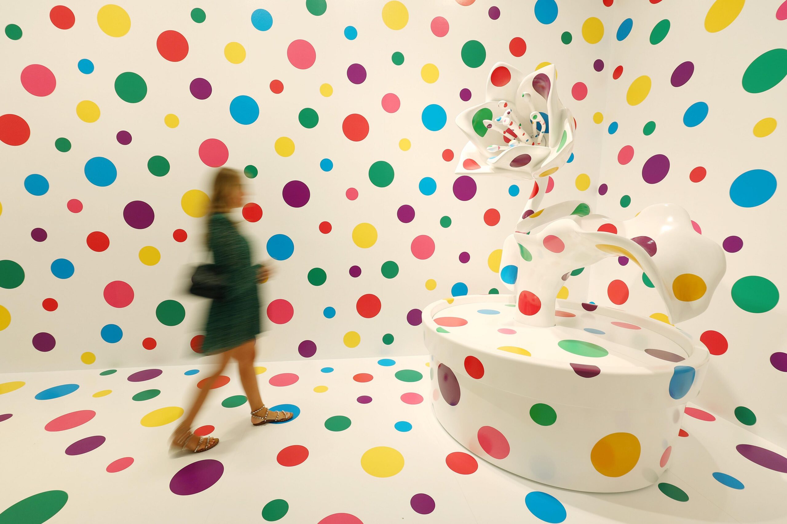 A blurred gallery visitor in front of a white wall and sculpture covered in multi-coloured spots