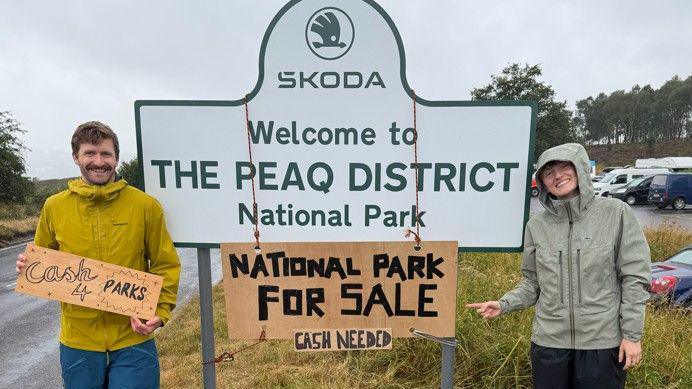 Two people at the side of a road sign which reads SKODA WELCOME TO THE PEAQ DISTRICT NATIONAL PARK. A cardboard sign is hung around the larger sign which reads NATIONAL PARK FOR SALE CASH NEEDED.