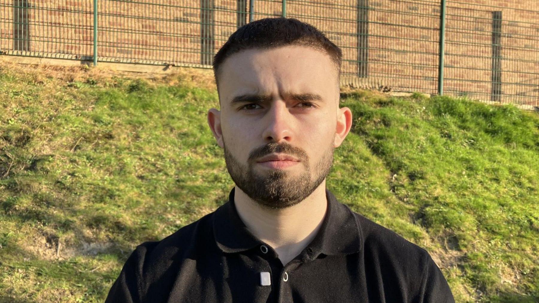 George is standing outside by some green playing fields and looking directly at the camera.  He has short dark hair and a neatly clipped dark beard. He has dark eyes and is wearing a black polo shirt.  He looks serious.
