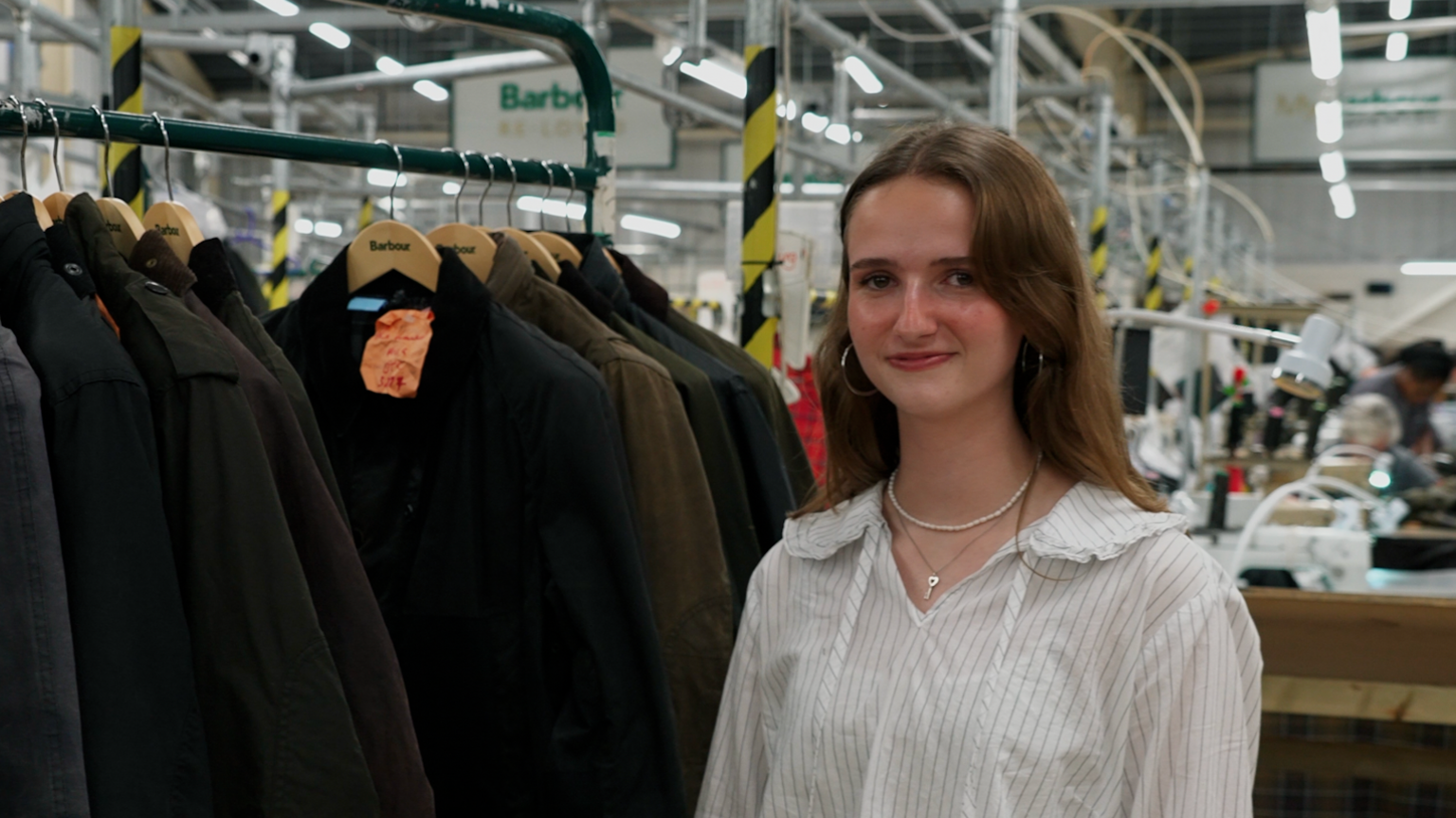 young woman standing in the foreground beside a clothing rail of dark waxed jackets on wooden Barbour hangers. The person is wearing a light striped shirt, hoop earrings and a necklace, and is facing the camera. The setting is a factory or workshop, with industrial lighting, metal framework and workstations visible in the background. Barbour signs can be seen above the clothing rail, and one jacket has an orange tag attached to it. No bags, bins or floor markers are clearly visible.