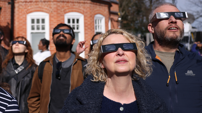 A group of people are standing in front of a red brick observatory. They are all wearing eclipse glasses with dark lenses and black cardboard frames.