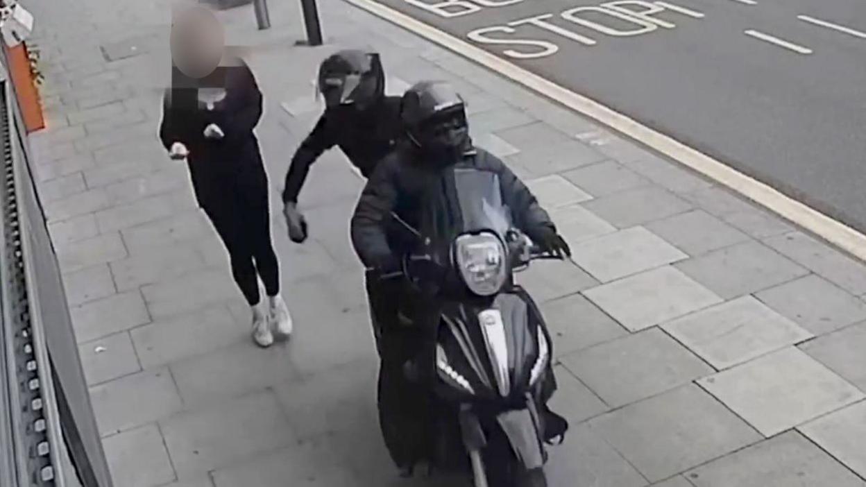 A CCTV image showing a black moped being driven along a pavement by two people wearing all black, snatching a mobile phone from a pedestrian walking along the pavement. One of the people on the moped can be seen holding the phone in their hand moments after snatching it, as the pedestrian recoils.