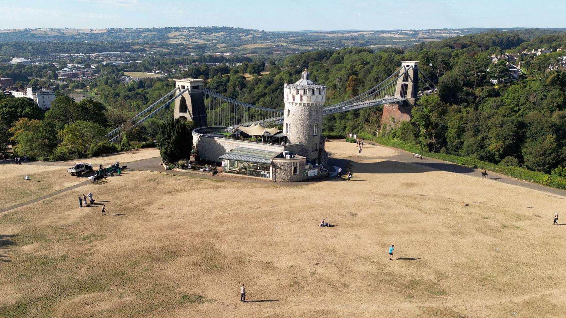 An aerial view shows Clifton Observatory next to Clifton suspension bridge west Bristol, with parched parkland around it with people walking on the brown grass, on 9 August.