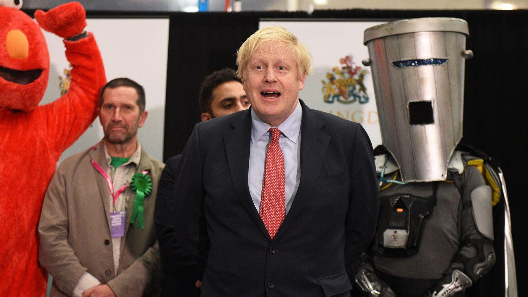 Boris Johnson is in the centre of the photo, wearing a black suit with a pale blue shirt and a red spotted tie. Behind him to the right is Count Binface, wearing a large silver bin on his head and a silver cape. On his right is a man dressed as the fuzzy red Sesame Street character Elmo.