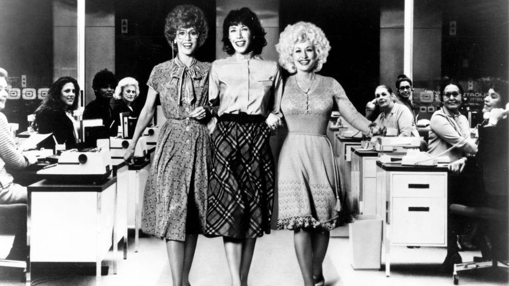 A black and white image shows a film still from 1980 comedy film 9 to 5, with Dolly Parton, her blond hair framing her face over a form-fitting dress, locking arms with actress Lily Tomlin, wearing a light-coloured blouse and dark-patterned skirt, and actress Jane Fonda, wearing a button-up dress with a scarf and glasses, as women sit at their desks looking on in an office
