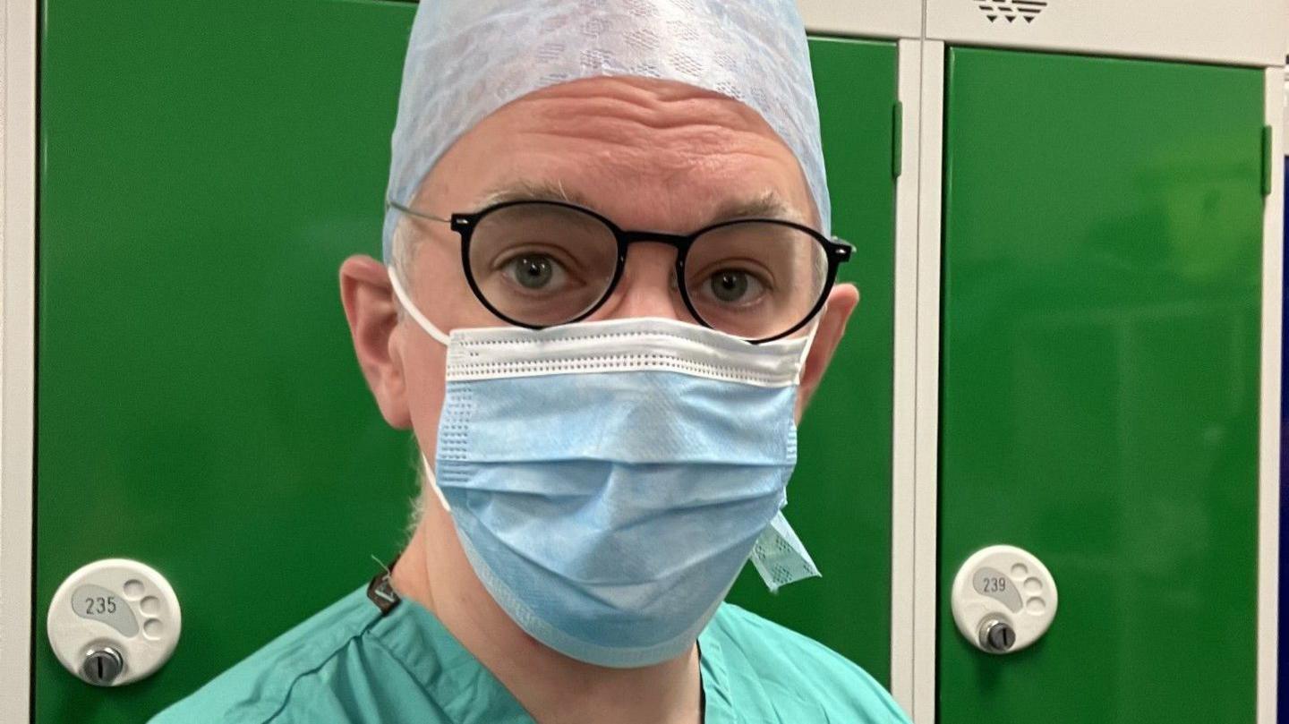 Head and shoulder selfie of correspondent James Gallagher, in green surgical scrubs, a blue facemask and a blue surgical hat. He is wearing dark blue glasses with metal arms. He is standing in front of a row of dark green lockers