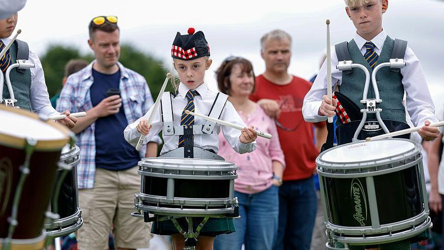 Two young drummers in traditional outfits playing drums that are strapped to them. There are spectators behind them