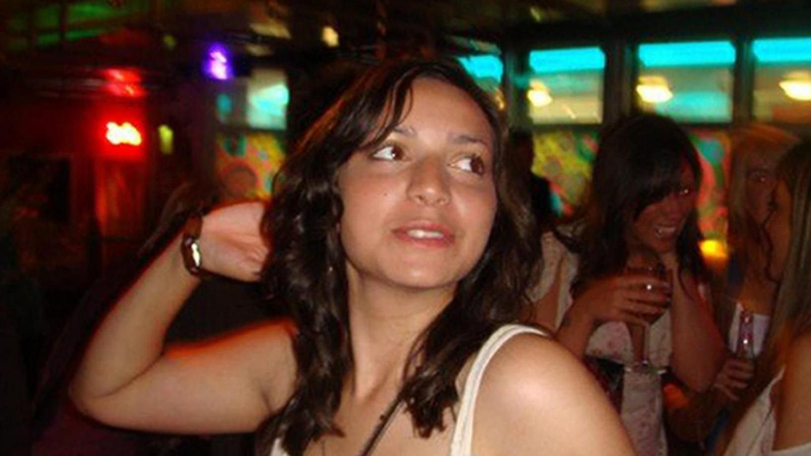Meredith Kercher. A young woman with shoulder‑length dark hair is pictured in a bar or club, turning slightly to her left with her arm raised near her head. She wears a light‑coloured sleeveless top. Behind her, several people are talking and holding drinks under colourful lighting, with red and turquoise tones visible in the background.