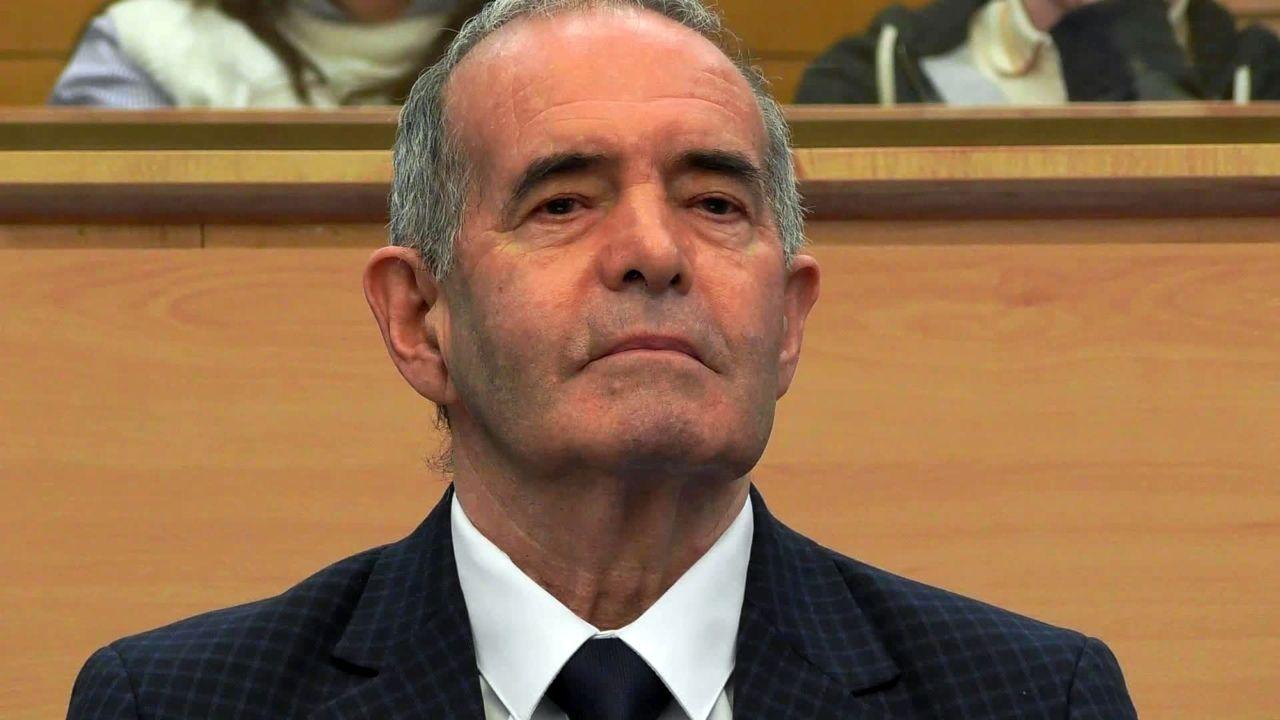 A smiling 77-year-old man in a blue suit and tie sits in the dock at the High Court in Glasgow