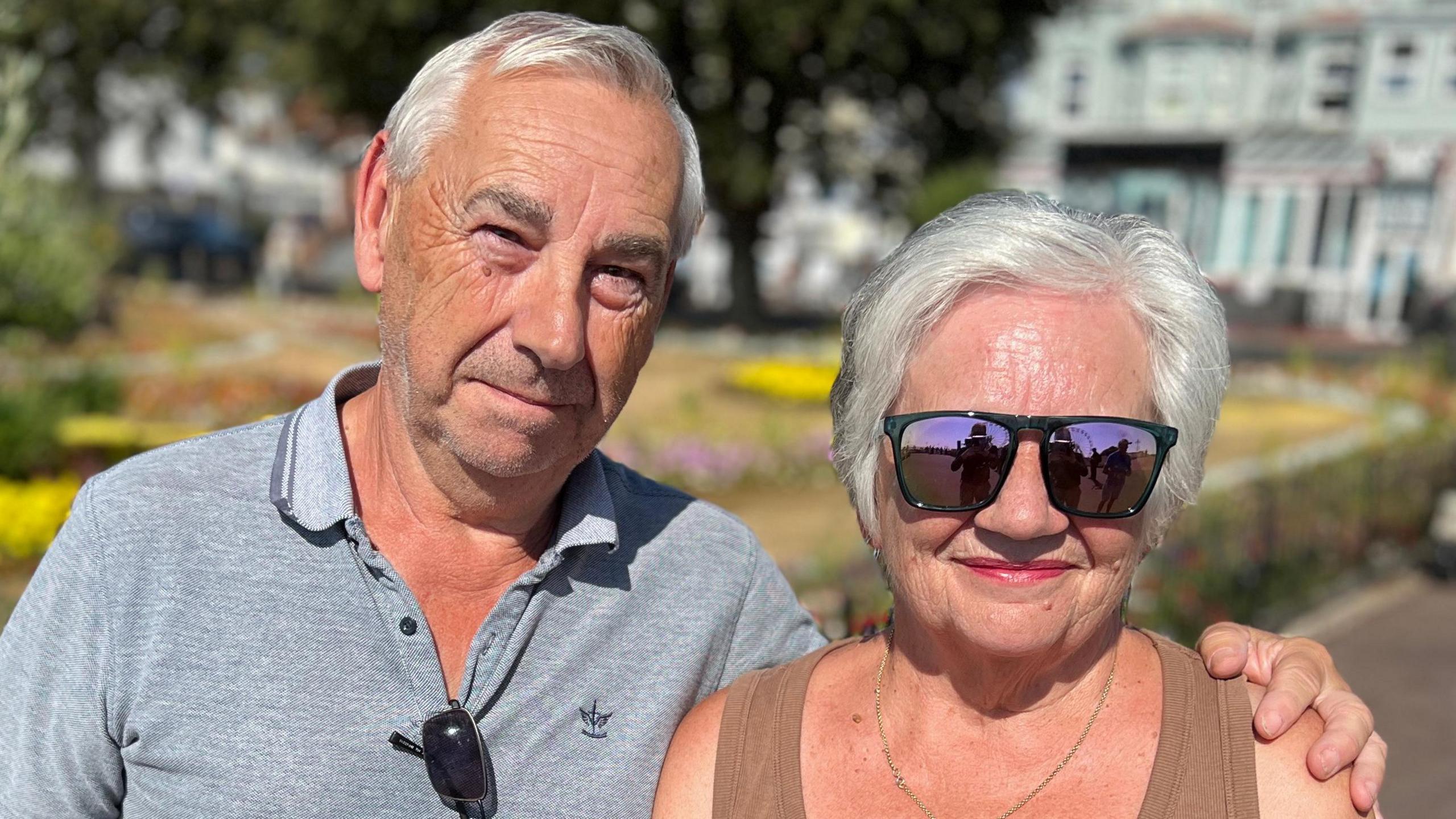 John and Sue Wackett standing by the seafront in Clacton. They both have grey hair. Sue is wearing a brown vest top and sunglasses. John is wearing a grey polo top and squinting due to the sun.