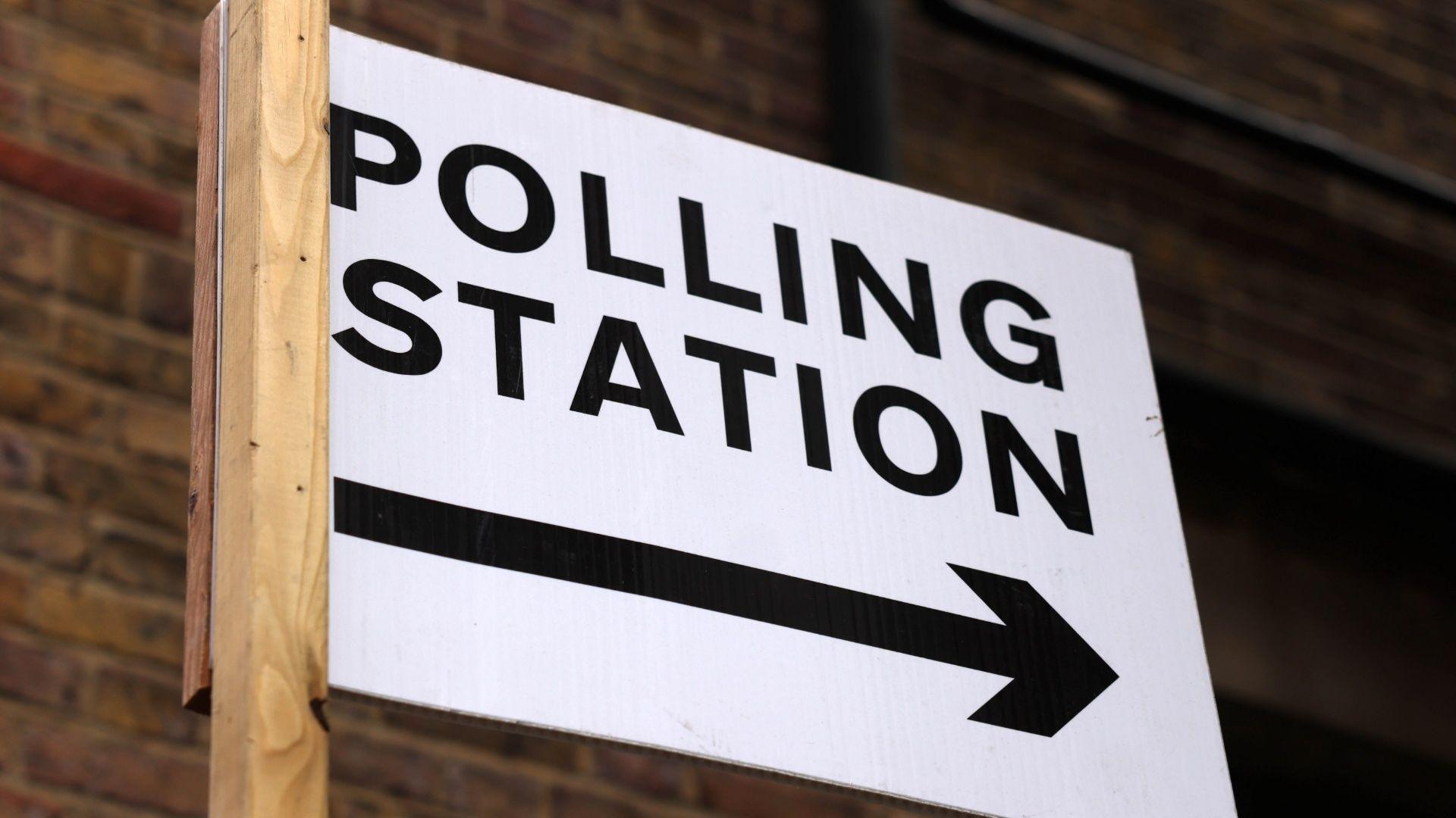 A white sign with black writing reading 'Polling Station' and a black arrow pointing left to right fixed to a wooden pole
