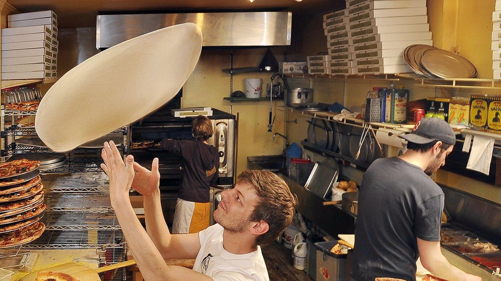 A chef tosses the base of a pizza into the air, behind him other kitchen staff work on pizzas.