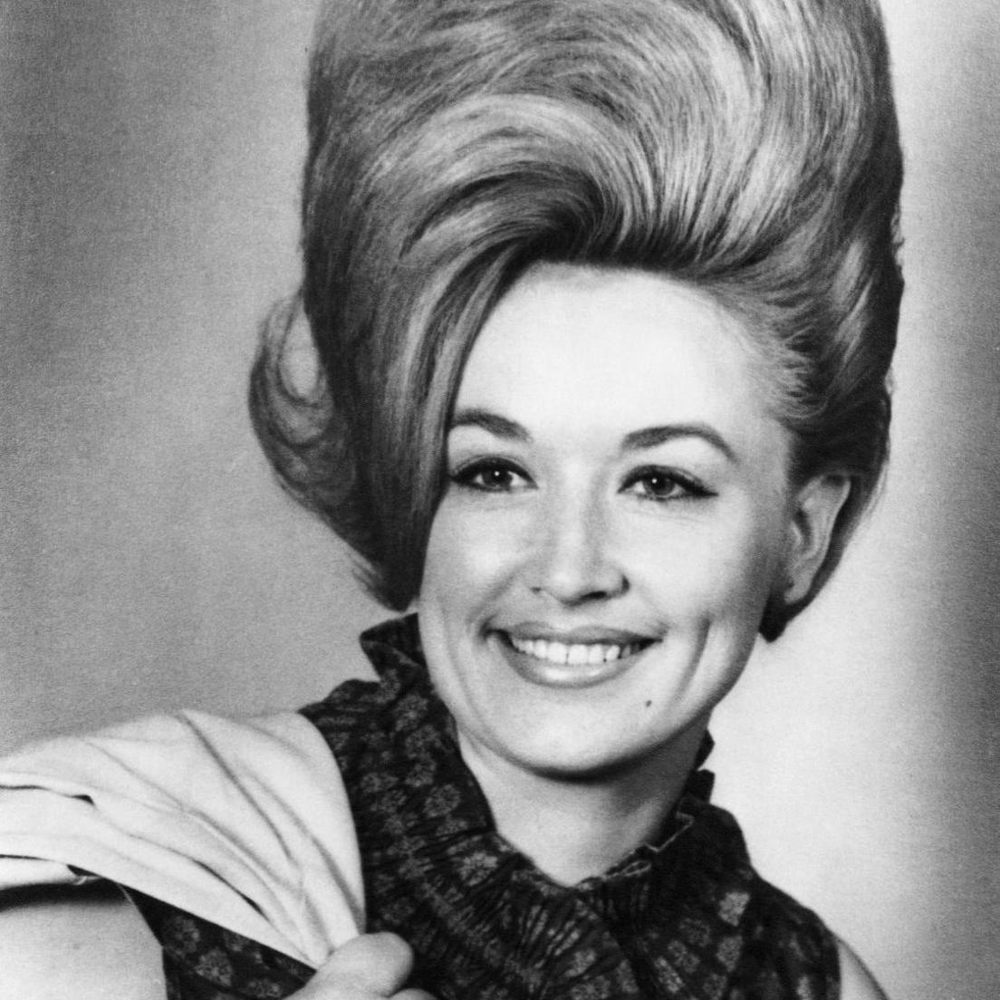 A black and white head shot of Dolly, smiling with a jacket slung over her shoulder. Her hair is nearly the same length as her face and stands back combed and high, with a curl leaning over to the left.