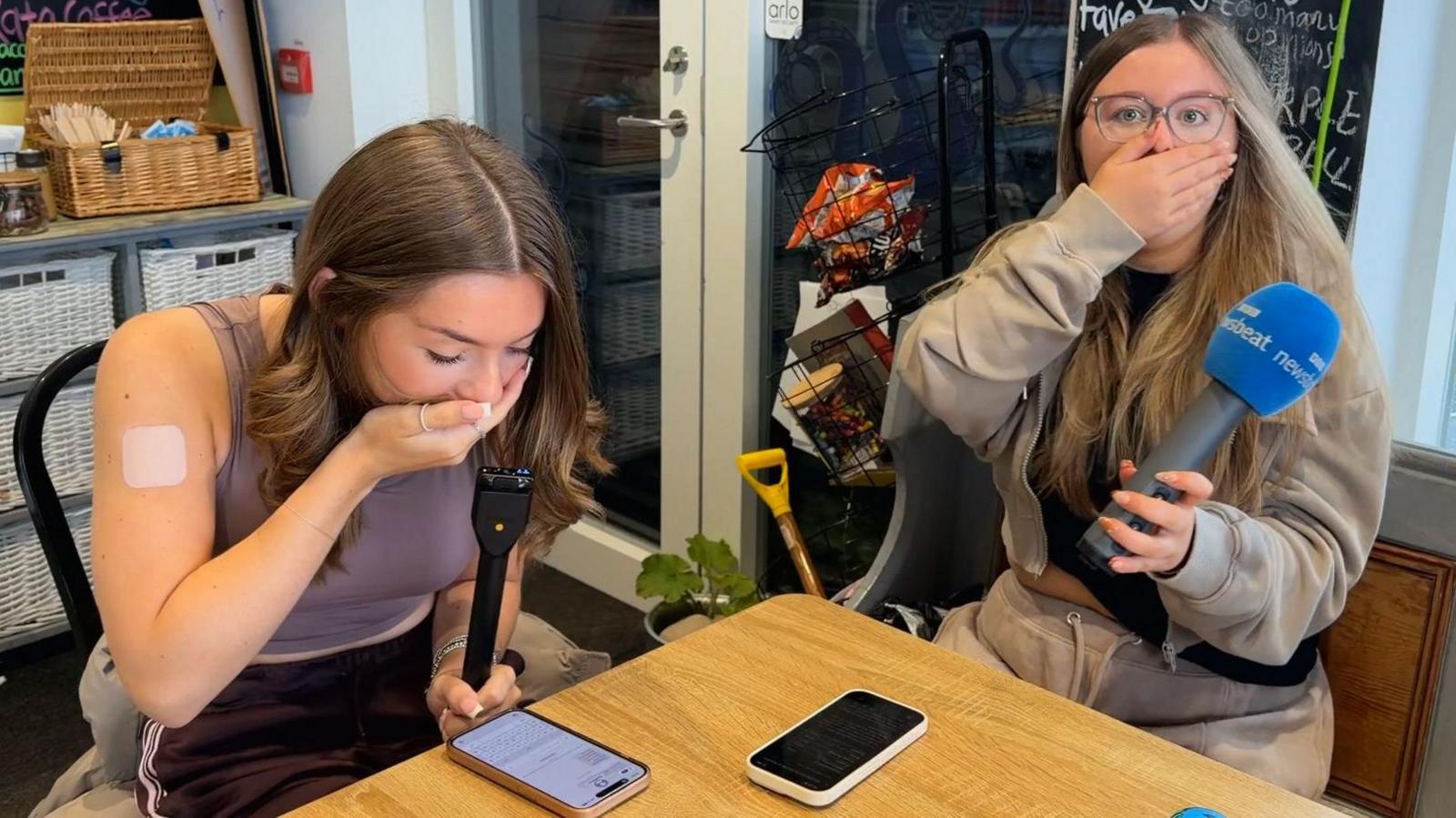 Two stunned girls react to their exam results with their hands over their mouths in shock. They both have long long, brown hair. Their phones are on a table. One is looking at her phone while the other, who is holding a microphone, is looking at the camera