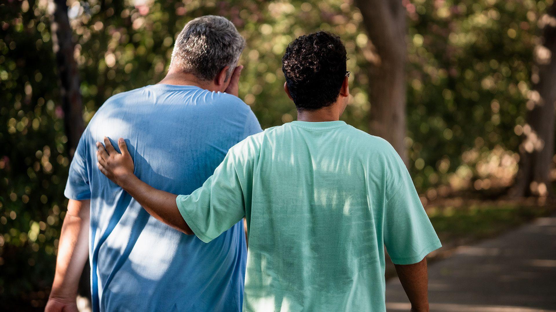 Stock photo shows the backs of two people walking through a shaded area, one is helping the other who appears to be struggling.
