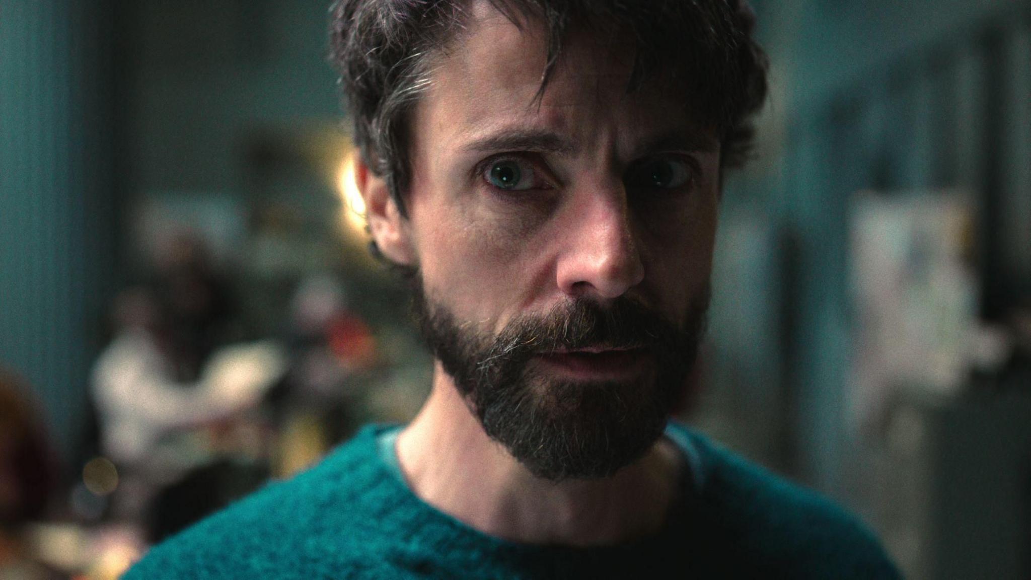 Close-up of Matthew Goode who is wearing a green jumper and is staring off to the right of the camera.