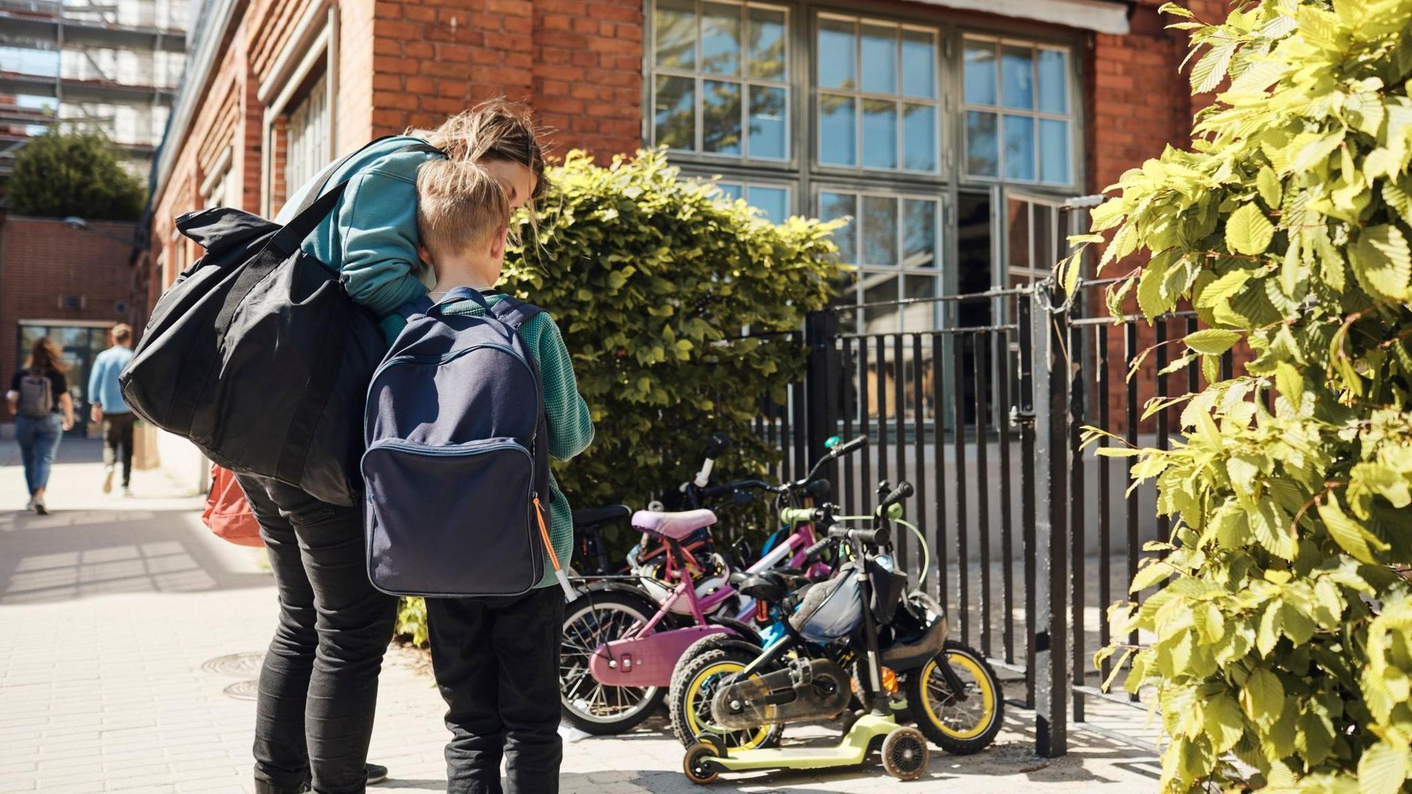 Stock image of a mum comforting a child outside a school. The boy has short blonde hair and a large blue backpack. The mum is wearing jeans and a teal jumper, and carrying a black shoulder bag. There are bikes and scooters outside the red-bricked school, and a black gate.