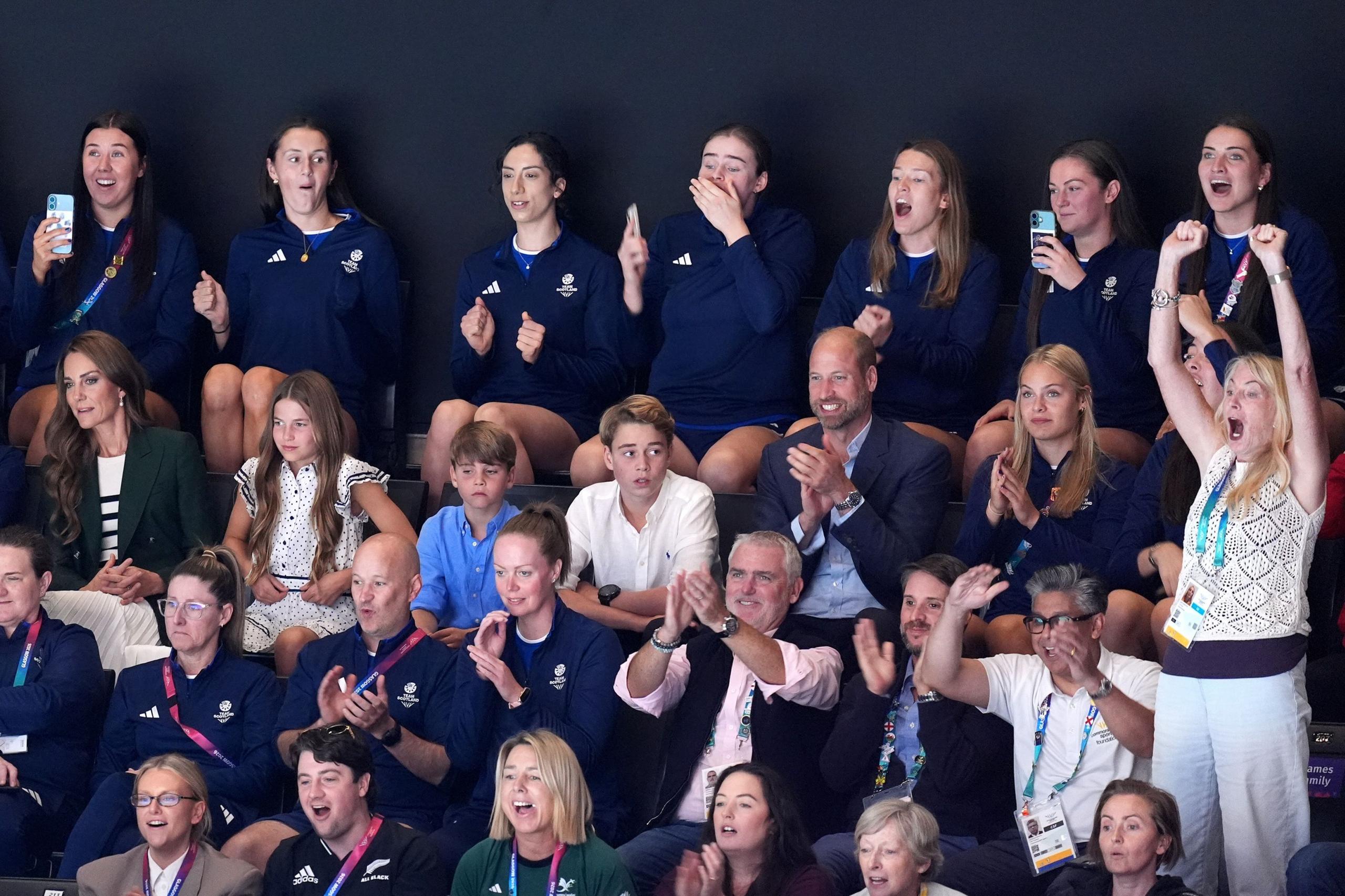 The Duke and Duchess of Rothesay with their three children watching a netball match. Everyone is seated while cheering, apart from a blonde woman on the far right of the photo who is standing with her arms in the air. She appears to be cheering loudly.