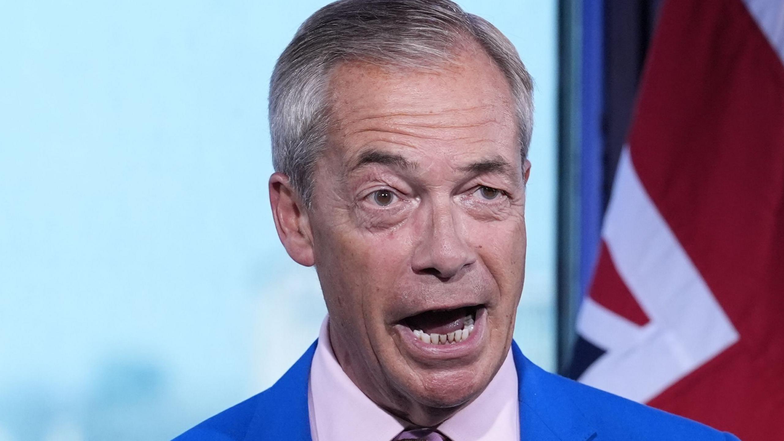 Nigel Farage, mid-way through making a statement. Only his head and shoulders are visible. He is animated, with his mouth open and one of his eyebrows is raised. Behind him on the right of the image is an out of focus Union Flag