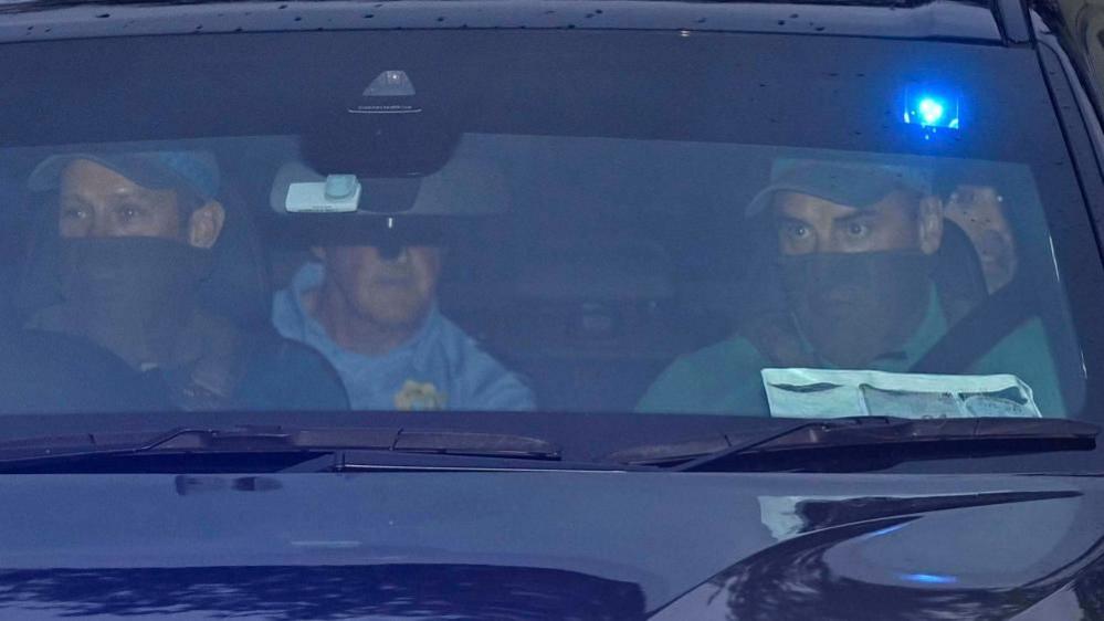 A close up image looking through the front windscreen of a vehicle. The two men in the front passenger seats are wearing baseball caps and have the bottom half of their face covered. There is a man on the left rear passenger seat wearing a blue shirt with a Gardai emblem. His face is obscured by the rear view mirror. On the right the face of Daniel Kinahan can be seen over the shoulder of one of the front passengers. He has stubble and is looking out the window.