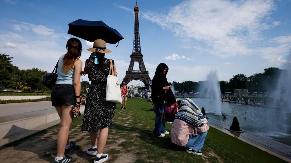 Women shelter from the heat under a black umbrella at the Trocadero Fountain by the Eiffel Tower, in Paris, France, 20 June 2026. Other people are seated next to the river.