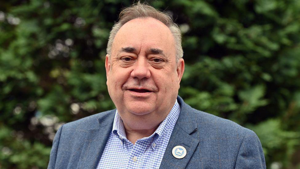 Alex Salmond in blue tweed jacket and blue and white checked open neck shirt. He is wearing a badge that cannot be identified. There are trees behind him.