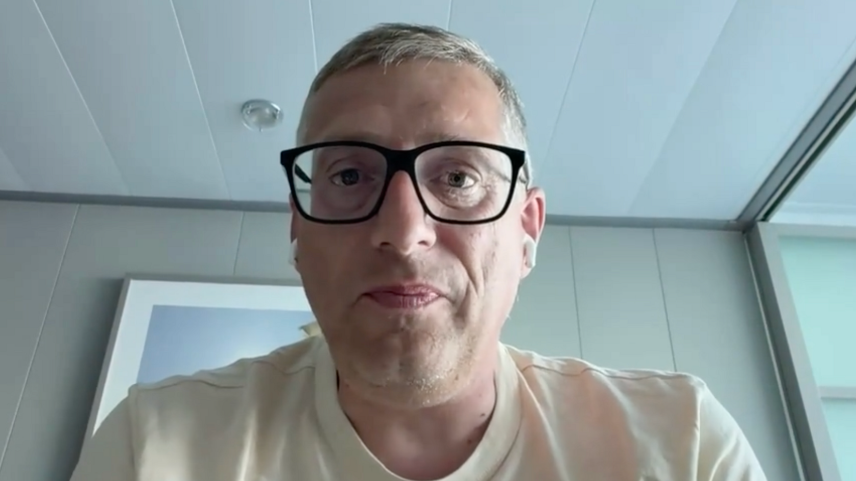 Simon Leyland has dark rimmed glasses, he is sitting in a cabin and wearing a beige tee-shirt