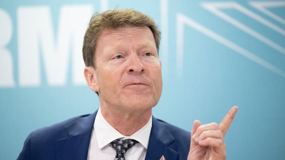 Richard Tice, in blue jacket, white shirt, and black and white tie, points with his finger while talking to reporters