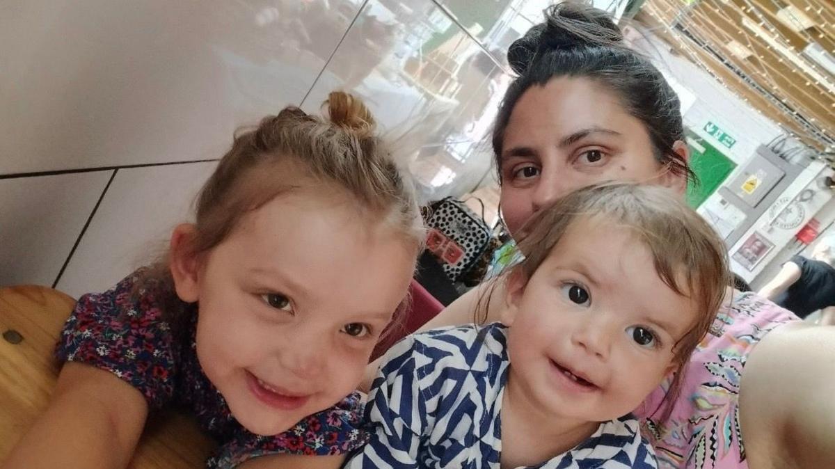 London Mother Harriet Anscombe, her brunette hair piled atop her head in a bun, holds her mobile away from her to take a selfie with her two daughters, aged four and two