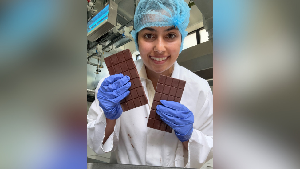 A woman holding two bars of chocolate while smiling into the camera. She is wearing a white gown with purple gloves and a blue hair net.