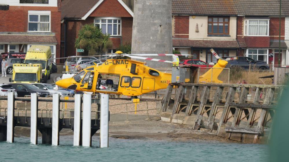 A yellow air ambulance helicopter is parked beside the waterfront near Shoreham Fort. Ambulance vehicles, parked cars and houses can be seen in the background.