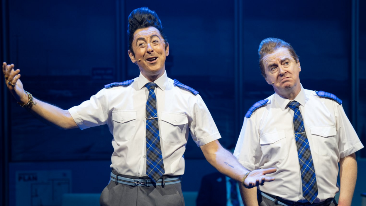 Two performers in matching airline-style uniforms perform on stage in The High Life: The Musical, gesturing as they sing under blue stage lighting.