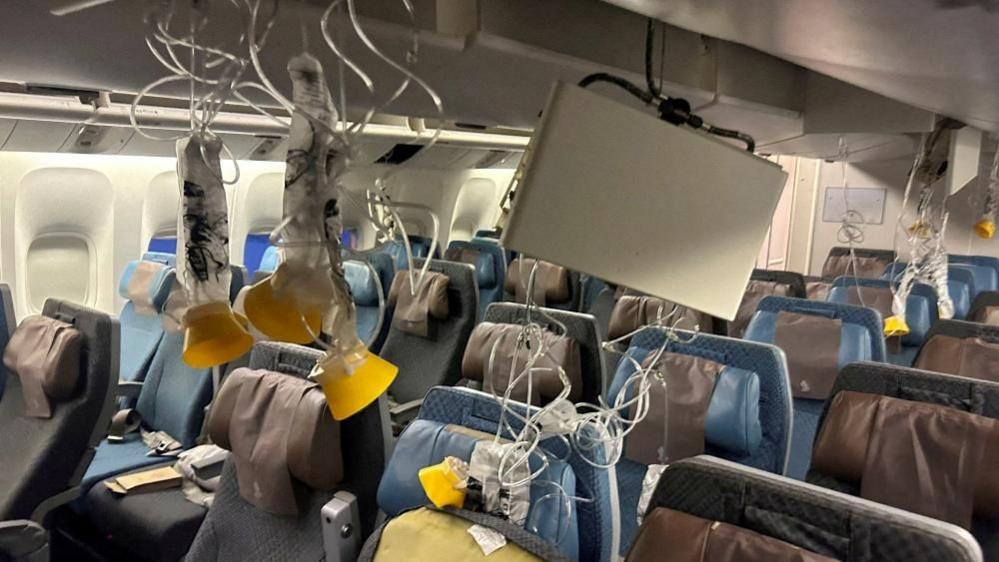 Oxygen masks hang from the ceiling inside an aircraft cabin. Rows of seats sit empty beneath them.