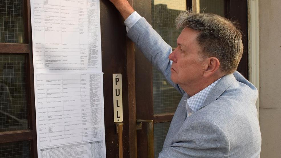 Ian Davidson posting the statement of persons nominated outside Clacton Town Hall. He is on the right, has short light brown hair and is wearing a light grey jacket over an open-neck shirt. His right arm is upraised as he puts the long list of typed names on to the brown wooden and glass door.