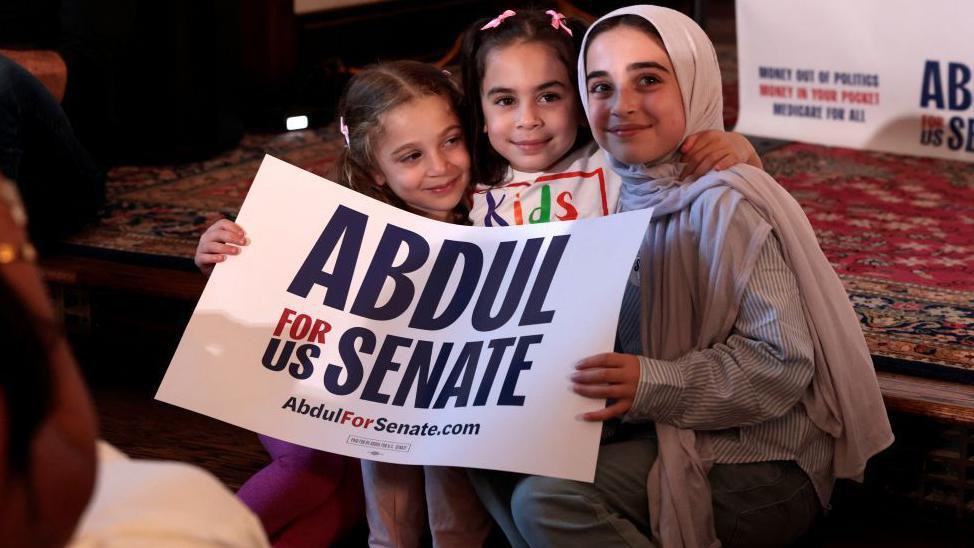 Three young girls, two with pink hair clips and one with a beige headscarf, smile and hold a sign saying "Abdul for US Senate".