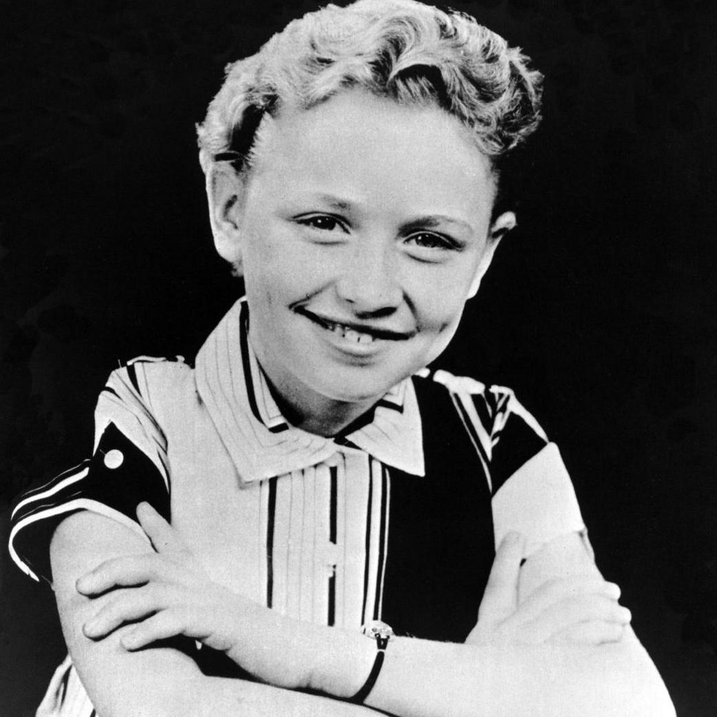 A black and white photo of Dolly as a child. She is sitting at a desk smiling and her hair is very short and curly.  She wears a conservative black and white striped short sleeved shirt buttoned up to her neck.