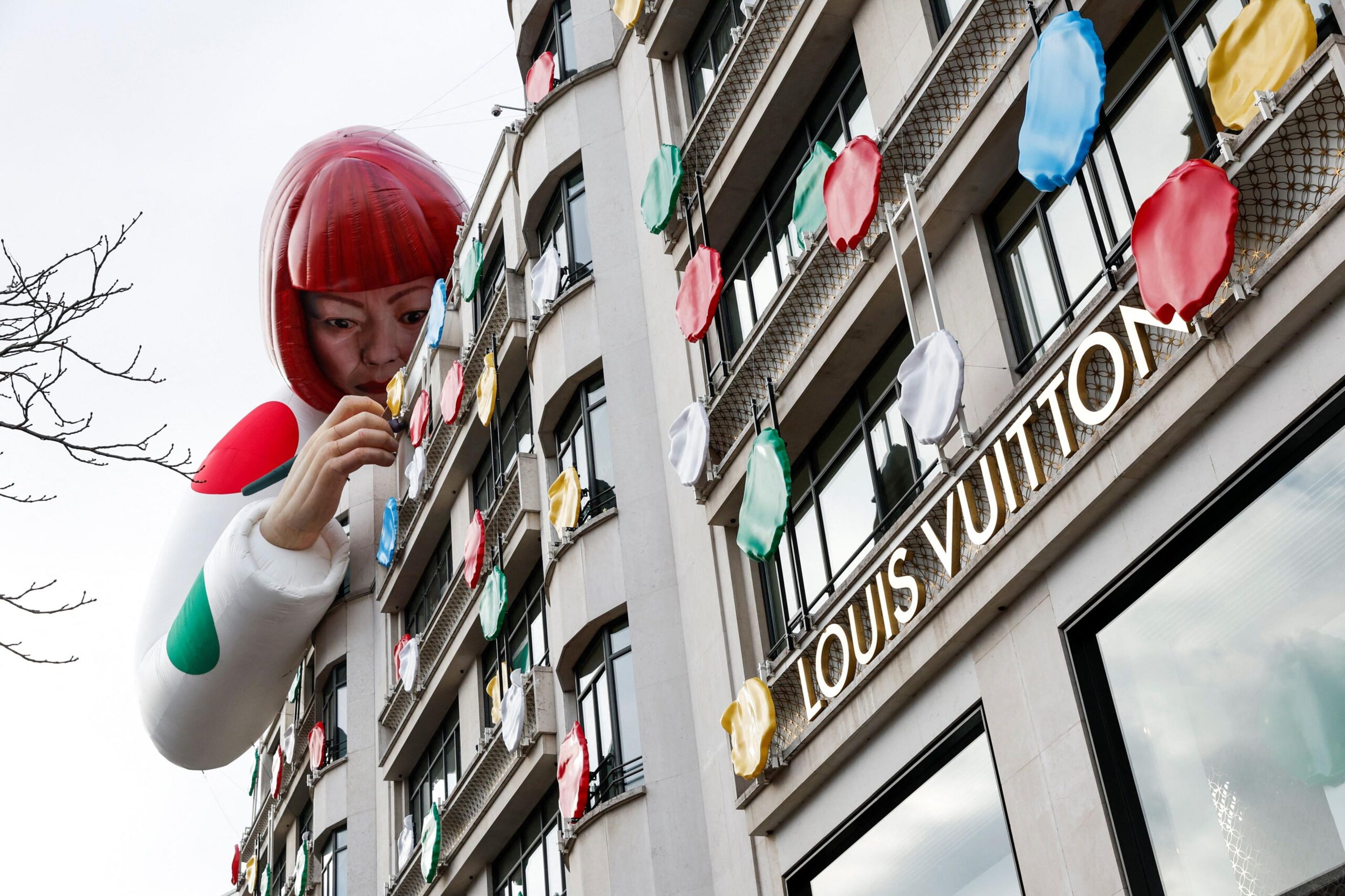 Exterior of Louis Vuitton store with logo and a giant artificial figure of Yayoi Kusama leaning over the roof and apparently attaching multi-coloured polka dots to the building