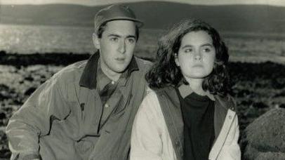 A black-and-white photograph shows Allan and Shirley seated among large rocks on a shoreline, with water and distant hills visible in the background. Both are dressed in outdoor clothing, creating a candid, natural scene in a rugged coastal setting.